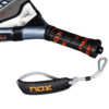 Eine Nahaufnahme des NOX AT10 Pro Cup Comfort 2025 Padelschlägergriffs zeigt einen schwarz-orangefarbenen Griff, der die Marke „Nox“ zeigt. Die abnehmbare Handschlaufe, die mit einem gepolsterten schwarzen Stück mit dem „Nox“-Logo verziert ist, ergänzt das metallisch-schwarze Design wunderbar.